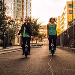 Two women riding INMOTION V5 unicycles on an urban street at sunset.